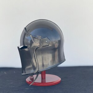 May include: A black helmet with a visor and a chainmail chin strap. The helmet is on a wooden stand.