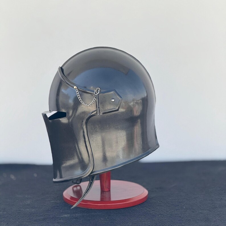 May include: A black helmet with a visor and a chainmail chin strap. The helmet is on a wooden stand.