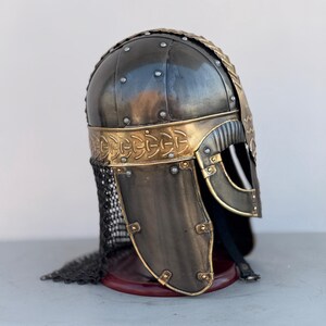 May include: A black and gold Viking helmet with a chainmail neck guard. The helmet has a decorative design on the top and a nose guard.