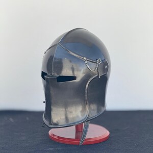May include: A black metal helmet with a visor and a chainmail chin strap. The helmet is on a red wooden stand.
