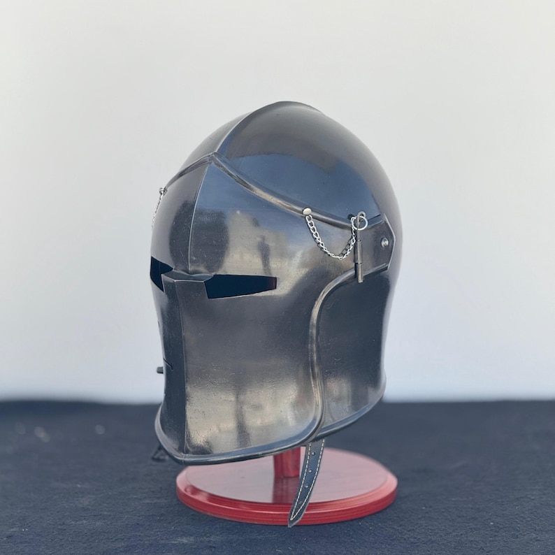 May include: A black metal helmet with a visor and a chainmail chin strap. The helmet is on a red wooden stand.