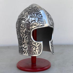May include: A silver helmet with intricate carvings on the surface. The helmet is on a red wooden stand.