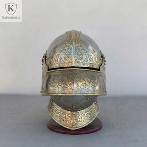 Op de afbeelding: Zilveren en gouden ridderhelm met ingewikkelde bloemmotieven. De helm wordt gepresenteerd op een houten standaard. Het logo van The Knights Ltd is linksboven zichtbaar.