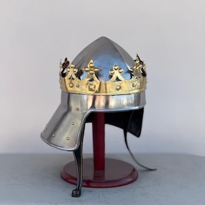 May include: A silver helmet with a gold crown, the crown has a pointed top and decorative details. The helmet is on a red stand.