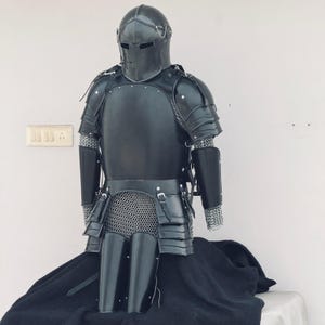 May include: A full suit of black armor with a helmet, chainmail, and plate armor. The armor is displayed on a black cloth background.