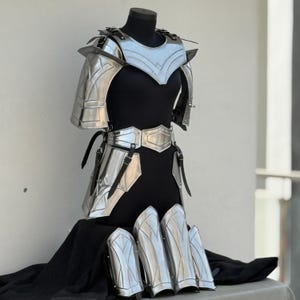 May include: A silver-colored metal armor set for the torso, arms, and legs. The armor is made up of plates and straps. The armor is displayed on a black fabric background.