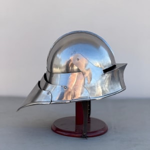 May include: A silver helmet with a pointed visor and a rounded top. The helmet is on a wooden stand.