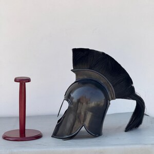 May include: A black Spartan helmet with a horsehair crest, displayed on a wooden stand. The helmet has a distinctive curved shape and a raised brow.