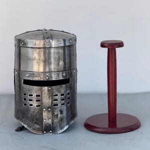 May include: A silver knight helmet with a visor and a decorative stand. The helmet has a textured surface and is made of metal. The stand is made of wood and is painted a dark red color.
