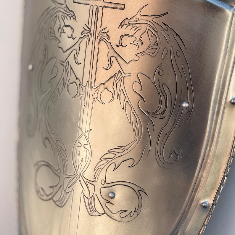 May include: A close-up of a metal shield with two dragons etched into the surface. The dragons are facing each other and have their wings spread out. The shield is a light gold color and has a smooth surface.