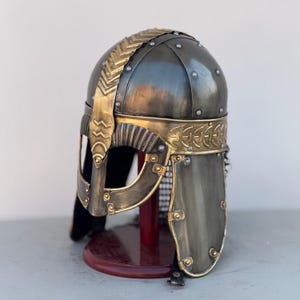 May include: A Viking helmet with a gold trim and a chainmail face mask. The helmet is made of metal and has a dark gray finish. It is displayed on a wooden stand.
