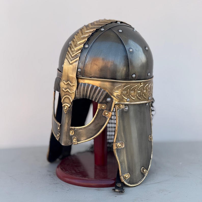 May include: A Viking helmet with a gold trim and a chainmail face mask. The helmet is made of metal and has a dark gray finish. It is displayed on a wooden stand.