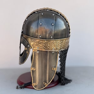 May include: A Viking helmet with a chainmail face mask. The helmet is made of metal and has a gold trim. The helmet is on a wooden stand.