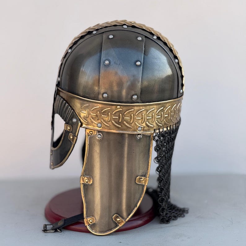 May include: A Viking helmet with a chainmail face mask. The helmet is made of metal and has a gold trim. The helmet is on a wooden stand.