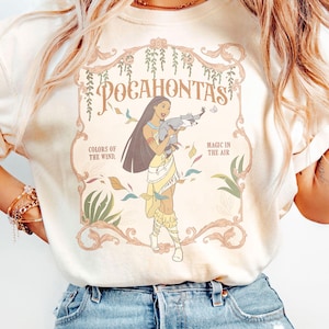 Op de afbeelding: Ivoor t-shirt met een afbeelding van Pocahontas die een wasbeer vasthoudt, met de tekst "Pocahontas" en "Colors of the Wind, Magic in the Air". Het ontwerp bevat bloemen- en bladeraccenten.