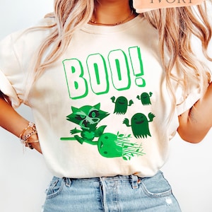 Könnte beinhalten: Elfenbeinfarbenes T-Shirt mit einem grünen Motiv eines Waschbären auf einem Besen, begleitet von drei grünen Geisterfiguren. Das Wort "BOO!" ist in großen grünen Buchstaben über dem Waschbären und den Geistern aufgedruckt. Ein lässiges Halloween-Design.