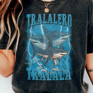 May include: A dark gray t-shirt with a graphic of three sharks wearing sneakers and the text "TRALALERO TRALALA" printed on the front.