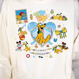 May include: A cream colored long-sleeved shirt with a colorful cartoon design featuring Mickey Mouse, Pluto, and other Disney characters. The design includes a heart with a blue background and a beach scene. The text "Summer 2023" is included in the design.
