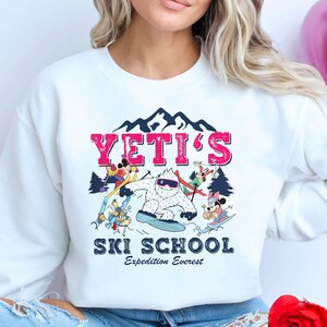 May include: White sweatshirt with a graphic of a Yeti and Mickey Mouse characters skiing. The text "YETI'S SKI SCHOOL Expedition Everest" is printed on the sweatshirt.