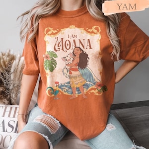 May include: Burnt orange t-shirt featuring a graphic of Moana holding a pig, with the text "I AM MOANA" in a decorative frame. The design includes tropical flowers, a rooster, and ocean waves. The shirt is a comfortable, casual style.