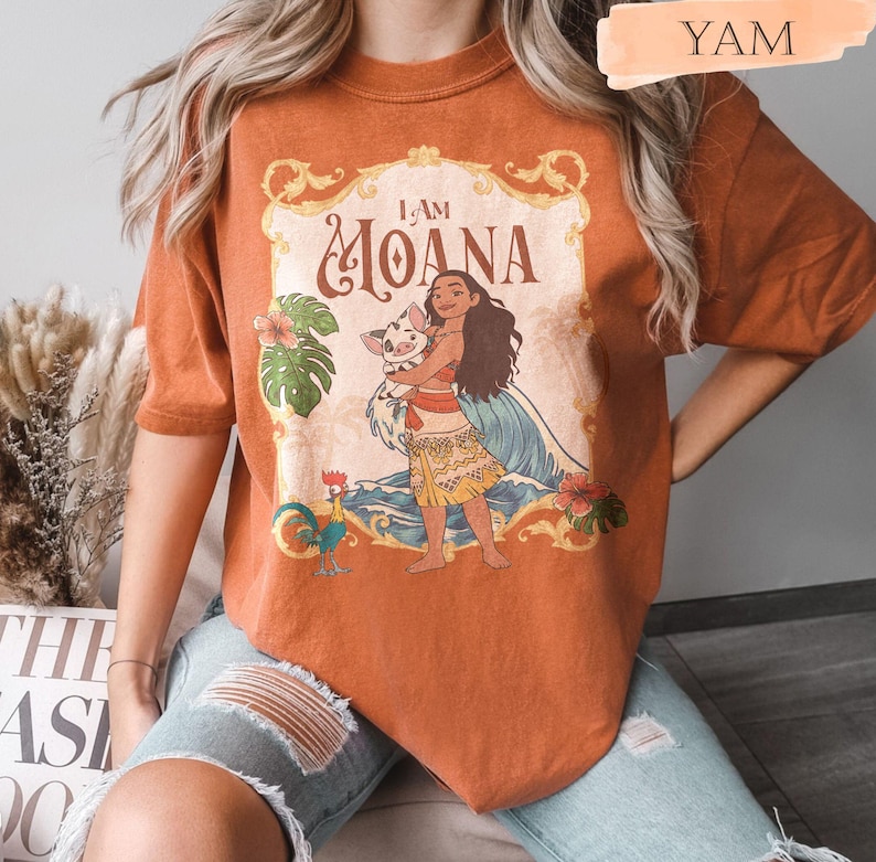May include: Burnt orange t-shirt featuring a graphic of Moana holding a pig, with the text "I AM MOANA" in a decorative frame. The design includes tropical flowers, a rooster, and ocean waves. The shirt is a comfortable, casual style.