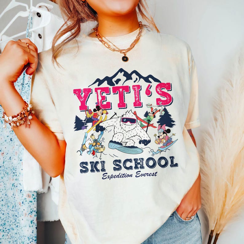 May include: A white t-shirt with a graphic of a yeti and cartoon characters skiing. The text "YETI'S SKI SCHOOL Expedition Everest" is printed on the shirt.