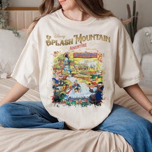May include: A white t-shirt with a colorful graphic print of the Disney Splash Mountain ride. The text "Disney Splash Mountain Adventure" is printed in gold on the shirt.