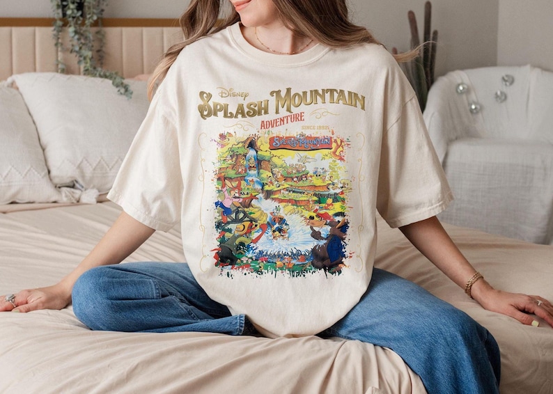 May include: A white t-shirt with a colorful graphic print of the Disney Splash Mountain ride. The text "Disney Splash Mountain Adventure" is printed in gold on the shirt.