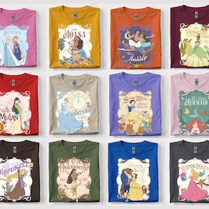 Op de afbeelding: Een verzameling opgevouwen t-shirts met verschillende Disney-prinsessenontwerpen. Elk shirt toont een ander personage of filmtitel, waaronder "Frozen", "Moana", "Aladdin" en "The Princess and the Frog". De shirts zijn in verschillende kleuren, zoals roze, oranje en blauw.