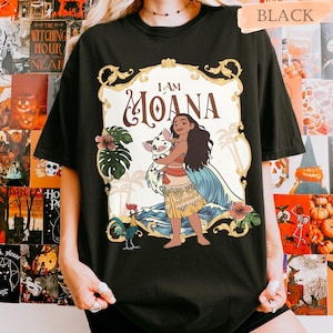 May include: Black t-shirt featuring a colorful graphic of Moana holding a pig, with the text "I AM MOANA". The design includes tropical elements like palm trees, flowers, and ocean waves. The shirt is a casual wear item.