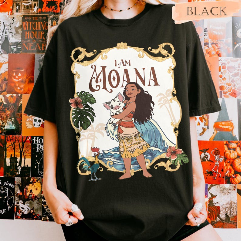 May include: Black t-shirt featuring a colorful graphic of Moana holding a pig, with the text "I AM MOANA". The design includes tropical elements like palm trees, flowers, and ocean waves. The shirt is a casual wear item.
