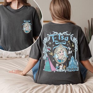 K&ouml;nnte beinhalten: Ein dunkelgraues T-Shirt mit einem Vintage-Motiv von Elsa aus Frozen. Das Motiv zeigt Elsa in einem blauen Kleid mit einer Krone und einem Schneeflocken-Design. Der Text "Elsa" ist in einer dekorativen Schriftart &uuml;ber ihr geschrieben. Der Text "Frozen Est. 2013" ist unter ihr geschrieben.