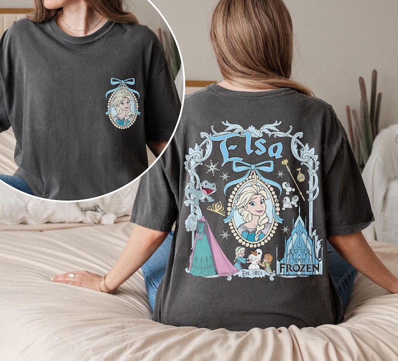 K&ouml;nnte beinhalten: Ein dunkelgraues T-Shirt mit einem Vintage-Motiv von Elsa aus Frozen. Das Motiv zeigt Elsa in einem blauen Kleid mit einer Krone und einem Schneeflocken-Design. Der Text "Elsa" ist in einer dekorativen Schriftart &uuml;ber ihr geschrieben. Der Text "Frozen Est. 2013" ist unter ihr geschrieben.