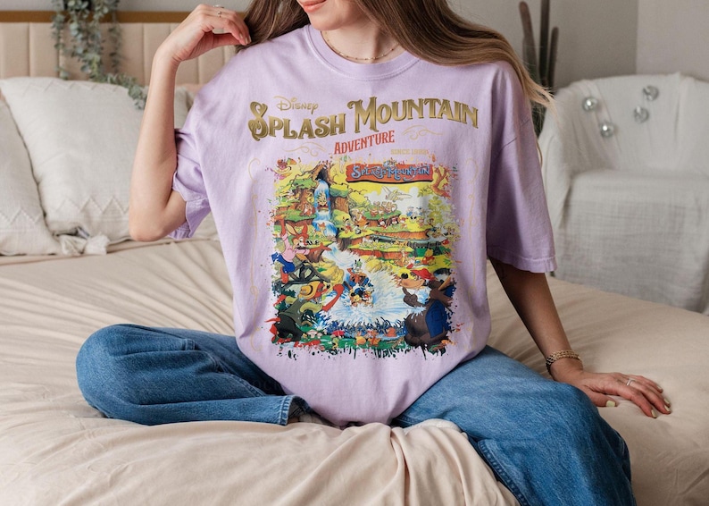 May include: A lavender t-shirt with a graphic print of a cartoon scene featuring the text "Disney Splash Mountain Adventure".