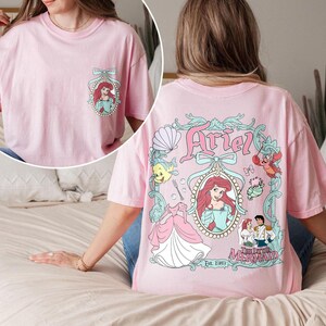 K&ouml;nnte beinhalten: Pinkes T-Shirt mit einem Aufdruck von Arielle aus Die kleine Meerjungfrau. Der Aufdruck zeigt Arielle in einem Muschelfass mit einer Krone und einer Schleife. Der Text "Ariel" befindet sich &uuml;ber dem Aufdruck und "Die kleine Meerjungfrau" unter dem Aufdruck. Der Text "Est 1989" befindet sich unter dem Aufdruck.