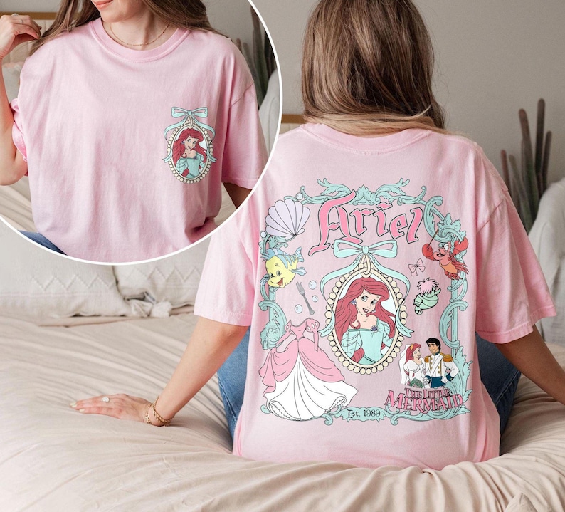 K&ouml;nnte beinhalten: Pinkes T-Shirt mit einem Aufdruck von Arielle aus Die kleine Meerjungfrau. Der Aufdruck zeigt Arielle in einem Muschelfass mit einer Krone und einer Schleife. Der Text "Ariel" befindet sich &uuml;ber dem Aufdruck und "Die kleine Meerjungfrau" unter dem Aufdruck. Der Text "Est 1989" befindet sich unter dem Aufdruck.
