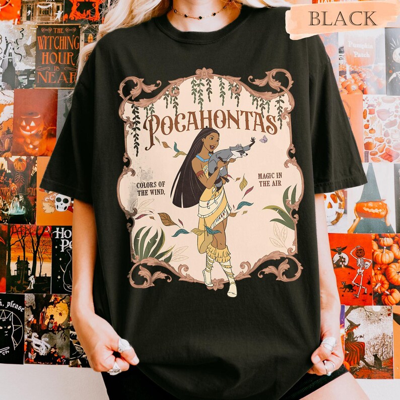 Op de afbeelding: Zwart T-shirt met een afbeelding van Pocahontas die een wasbeer vasthoudt. Het ontwerp bevat de tekst "Pocahontas", "Colors of the Wind" en "Magic in the Air", omlijst door decoratieve krullen en bladeren.