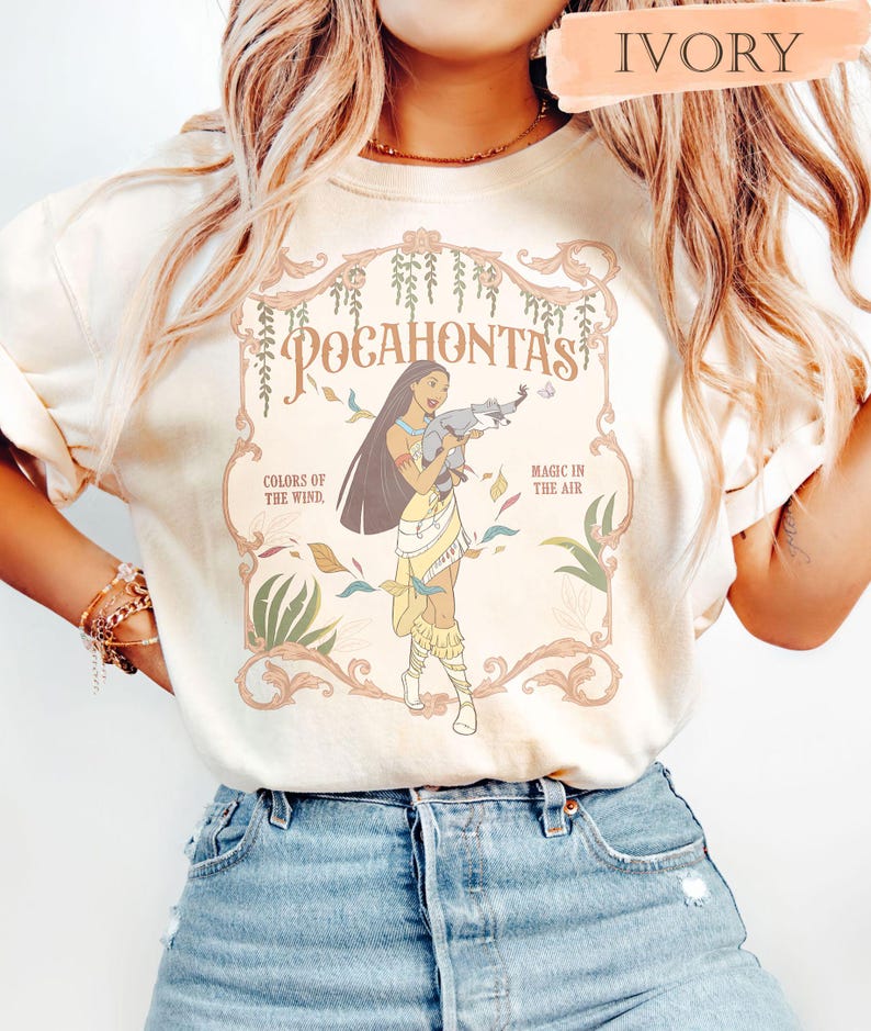 Op de afbeelding: Ivoor t-shirt met een afbeelding van Pocahontas die een wasbeer vasthoudt, met de tekst "Pocahontas" en "Colors of the Wind, Magic in the Air". Het ontwerp bevat bloemen- en bladeraccenten.
