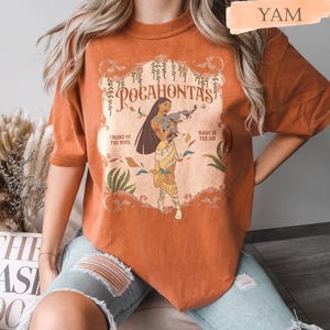 Op de afbeelding: Verbrand oranje t-shirt met een afbeelding van Pocahontas en het woord "Pocahontas". Het ontwerp bevat de zinnen "Colors of the Wind" en "Magic is in the Air". Het shirt heeft korte mouwen en een ronde hals.