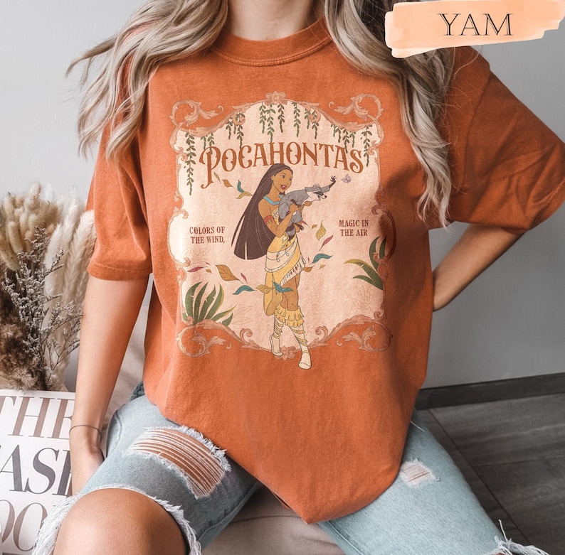 Op de afbeelding: Verbrand oranje t-shirt met een afbeelding van Pocahontas en het woord "Pocahontas". Het ontwerp bevat de zinnen "Colors of the Wind" en "Magic is in the Air". Het shirt heeft korte mouwen en een ronde hals.
