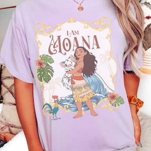 May include: A light purple t-shirt featuring a graphic of Moana holding a pig, with the text "I AM MOANA" in a decorative frame. The design includes tropical flowers, a rooster, and ocean waves.