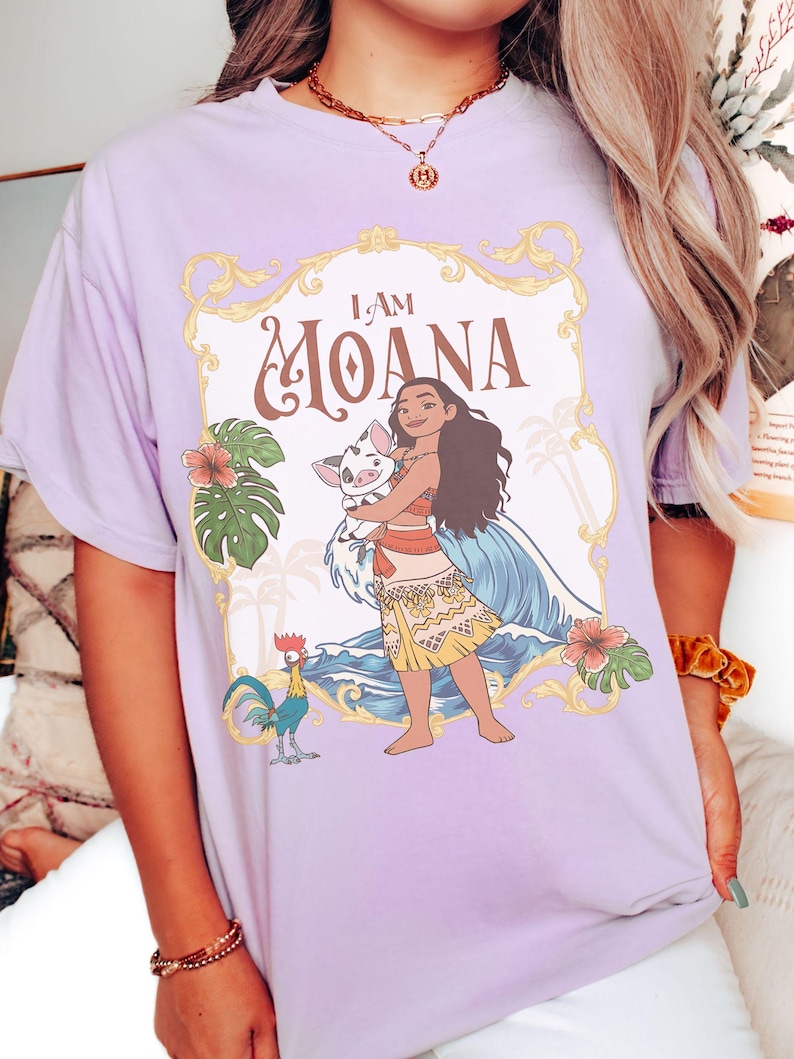 May include: A light purple t-shirt featuring a graphic of Moana holding a pig, with the text "I AM MOANA" in a decorative frame. The design includes tropical flowers, a rooster, and ocean waves.