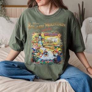 May include: A green t-shirt with a vintage-style graphic of the Splash Mountain ride at Disney World. The graphic features a colorful illustration of the ride's characters and scenery. The text "Disney Splash Mountain" is printed in gold lettering.