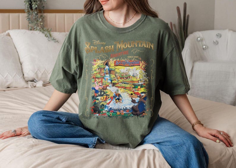 May include: A green t-shirt with a vintage-style graphic of the Splash Mountain ride at Disney World. The graphic features a colorful illustration of the ride's characters and scenery. The text "Disney Splash Mountain" is printed in gold lettering.