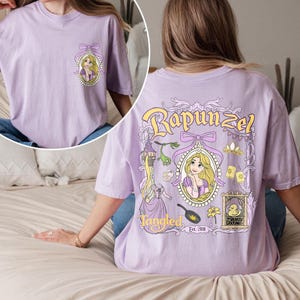 K&ouml;nnte beinhalten: Ein lavendelfarbenes T-Shirt mit einem Grafikdruck von Rapunzel aus dem Disney-Film Rapunzel - Neu verf&ouml;hnt. Die Grafik enth&auml;lt Rapunzels Portr&auml;t, einen Turm, eine Bratpfanne, eine Blume und eine Ente. Der Text "Tangled Est. 2010" ist unter der Grafik gedruckt.
