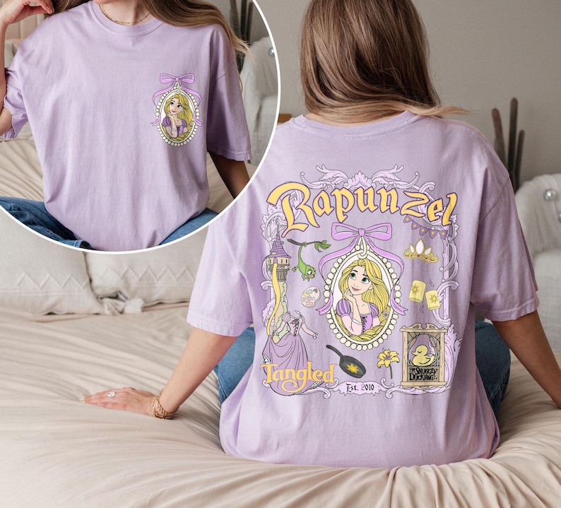 K&ouml;nnte beinhalten: Ein lavendelfarbenes T-Shirt mit einem Grafikdruck von Rapunzel aus dem Disney-Film Rapunzel - Neu verf&ouml;hnt. Die Grafik enth&auml;lt Rapunzels Portr&auml;t, einen Turm, eine Bratpfanne, eine Blume und eine Ente. Der Text "Tangled Est. 2010" ist unter der Grafik gedruckt.