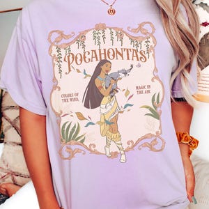 Op de afbeelding: Lichtpaars T-shirt met een Pocahontas-afbeelding. Het ontwerp bevat de tekst "Pocahontas", "Colors of the Wind" en "Magic in the Air". De illustratie toont Pocahontas met lang zwart haar, gekleed in een gele jurk en met een wasbeer.