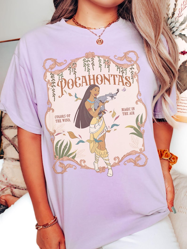 Op de afbeelding: Lichtpaars T-shirt met een Pocahontas-afbeelding. Het ontwerp bevat de tekst "Pocahontas", "Colors of the Wind" en "Magic in the Air". De illustratie toont Pocahontas met lang zwart haar, gekleed in een gele jurk en met een wasbeer.
