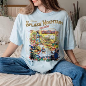 May include: A light blue t-shirt with a graphic print of the Splash Mountain ride at Disney World. The graphic features a colorful illustration of the ride's characters and scenery. The text "Disney Splash Mountain Adventure:  The  Splash-tacular" is printed on the shirt.