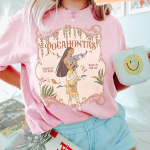 Op de afbeelding: Zachtroze t-shirt met een Pocahontas-afbeelding en de tekst "Pocahontas, Kleuren van de Wind, Magie in de Lucht". Het shirt wordt gedragen met een blauwe short en een witte mok met een gele smiley.